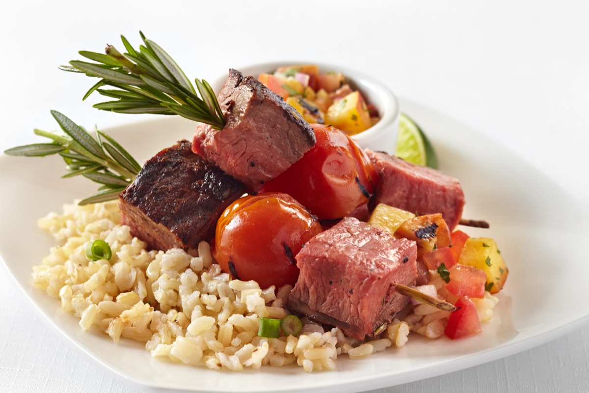 A white plate of grilled steak kabob with blistered tomatoes, brown rice, peach salsa and rosemary skewers
