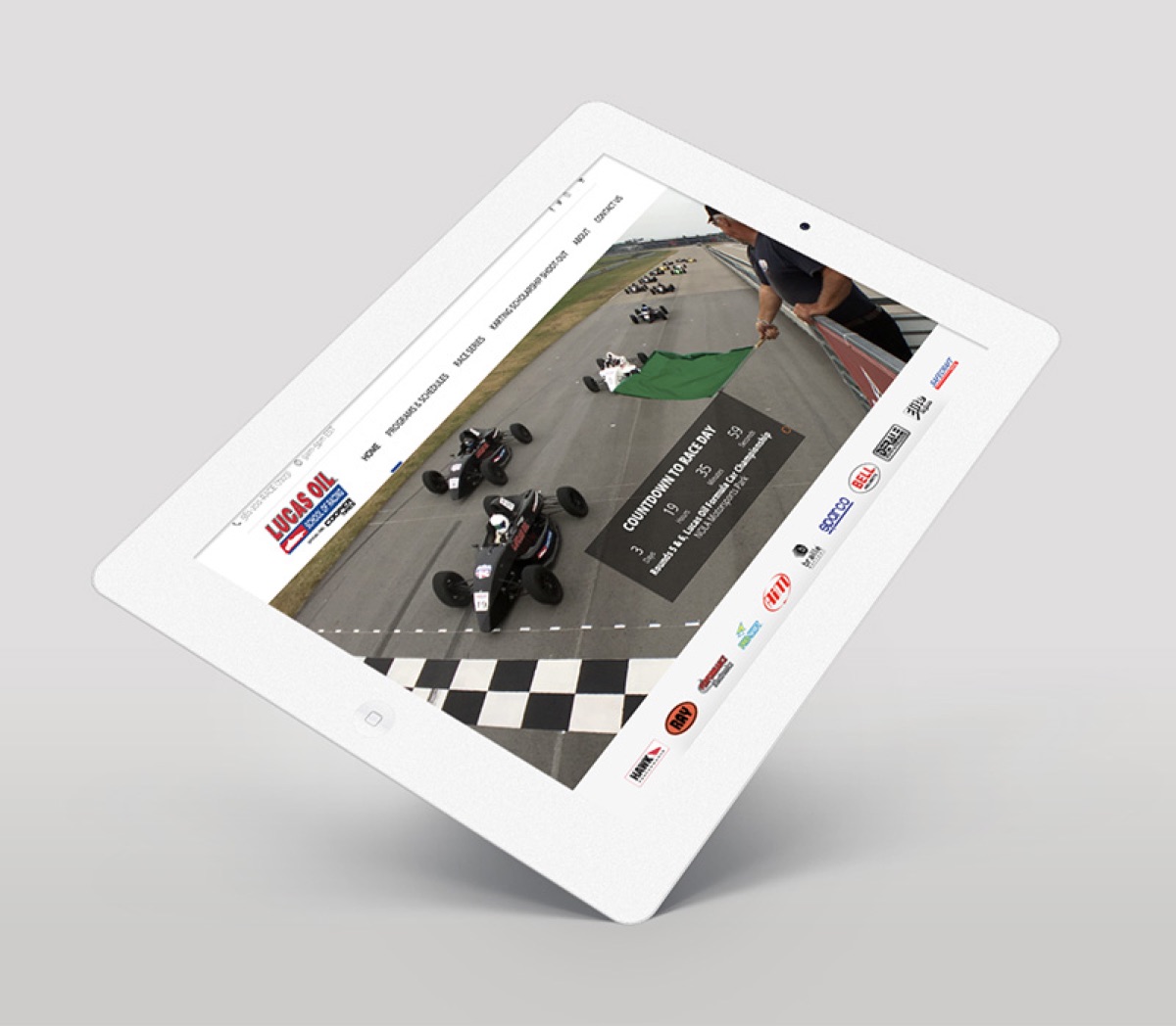 The Lucas Oil School of Racing website shown on a tilted white tablet, open on a race-day countdown over a photograph of open-wheel cars at a start line